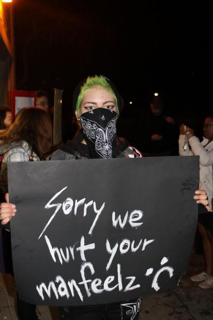 A masked protestor at the University of Toronto holding a sign that says "sorry we hurt your man feels."