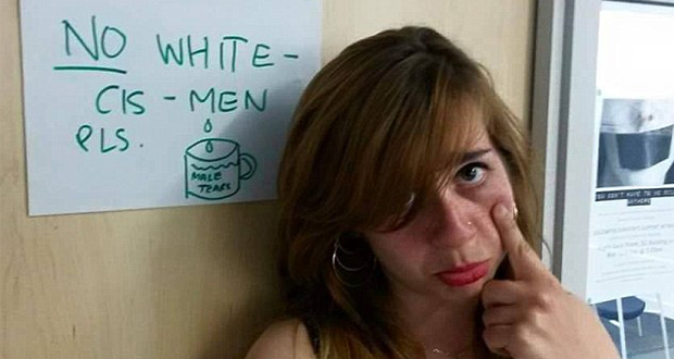 A photo of Bahar Mustafa mockingly pretending to shed tears. She stands next to a door with a sign that reads "No white cis-men please" above a cup that reads "male tears."